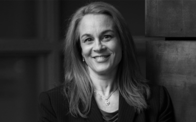 BW FacultyPhotos Lori Rosenkopf A black and white headshot of a smiling person with long hair, wearing a suit jacket and a necklace.