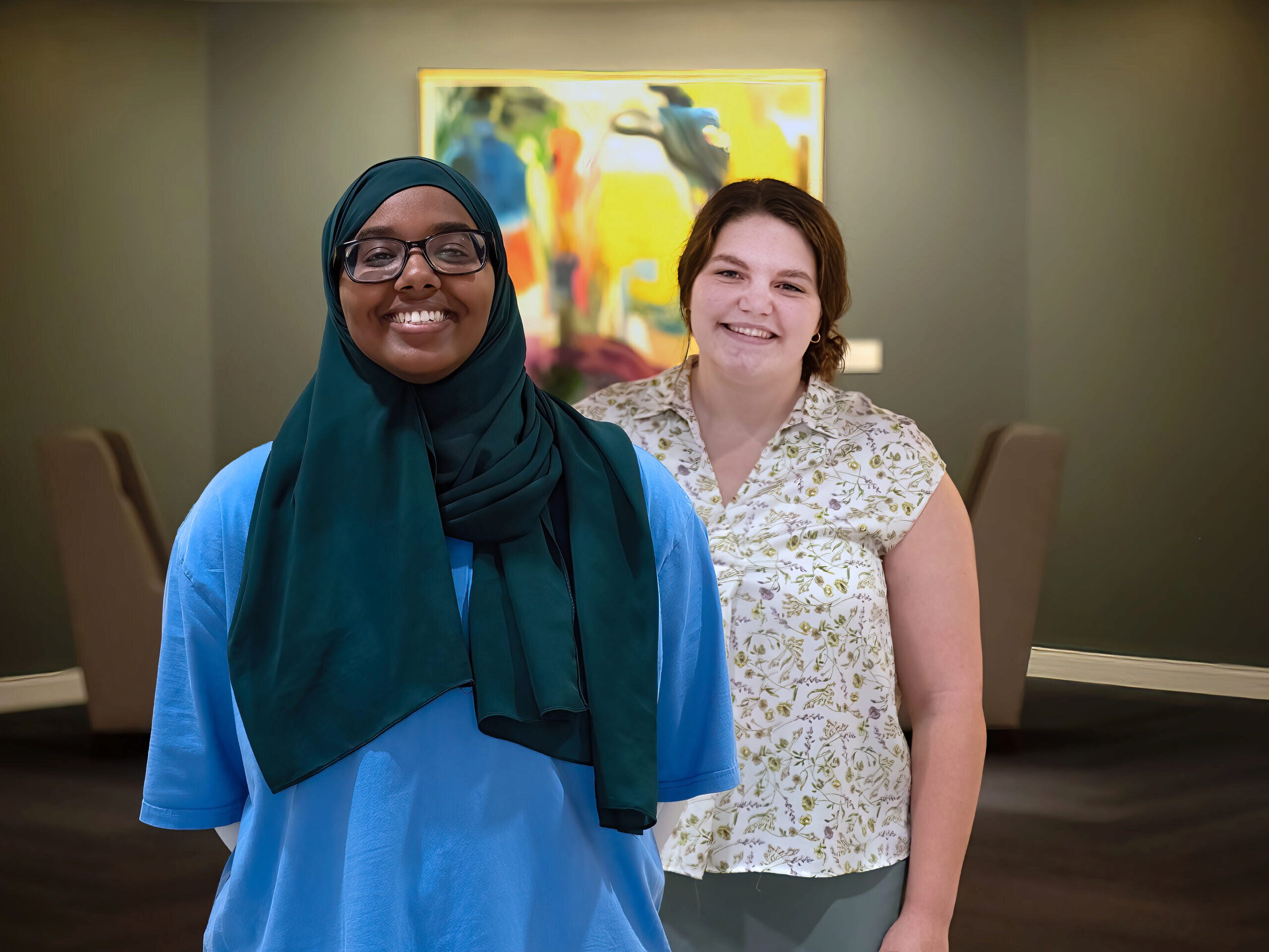 P9069461 (1) Two smiling people standing in a room with a colorful abstract painting on the wall behind them. One is wearing a hijab and glasses, the other has a floral-patterned top.