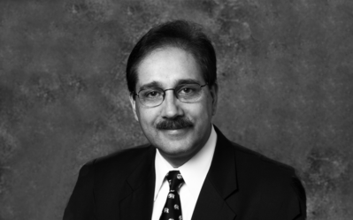 BW_FacultyPhotos-Harbir-Singh Headshot of a person wearing glasses, a suit, and a patterned tie against a textured background.