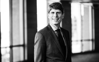 BW FacultyPhotos Nicolaj Siggelkow Black and white headshot of a person in a suit and tie, smiling, standing in a well-lit hallway with large windows.